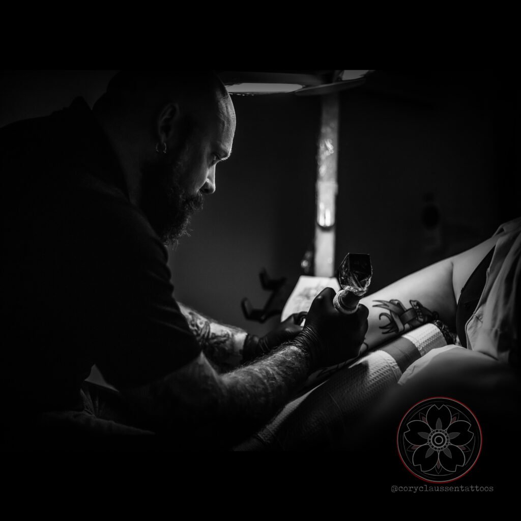 Cory Claussen, head of Red Arbor Tattoo, focused on tattooing a client’s arm in a dimly lit studio.