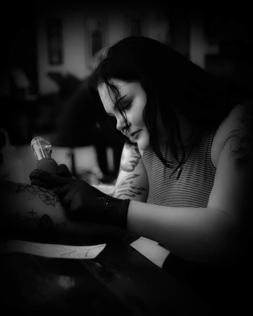 Savannah Layne tattooing a client with focused attention in a black-and-white photograph, showcasing her artistry and precision.