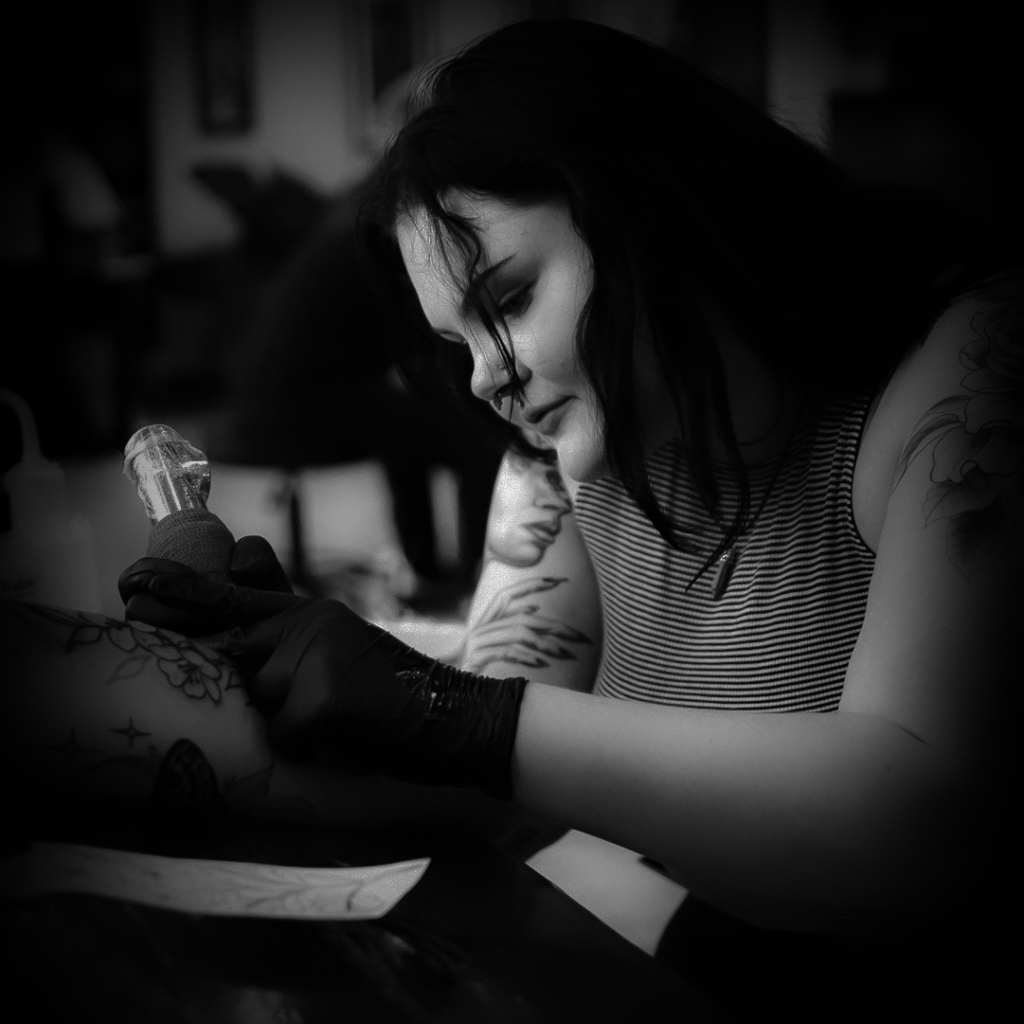 Savannah Layne tattooing a client with focused attention in a black-and-white photograph, showcasing her artistry and precision.