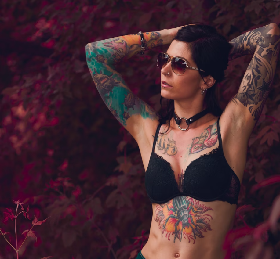 A woman showcasing vibrant tattoo sleeves, chest, and stomach tattoos, posing confidently outdoors against a backdrop of deep red foliage.