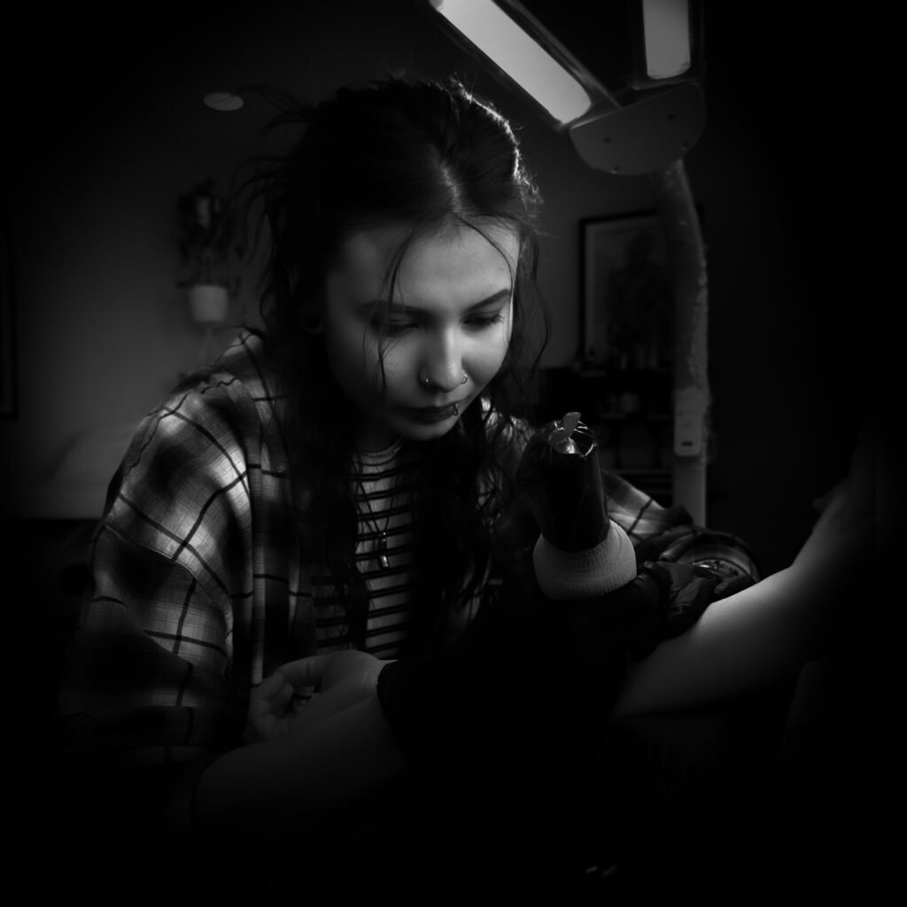 Mia Vrooman focused intently on tattooing a client, captured in a black-and-white photograph under soft studio lighting.